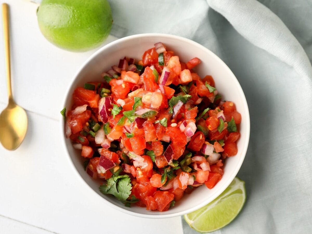a bowl of pico de gallo tomato salsa garnished with fresh herbs and a side of lime